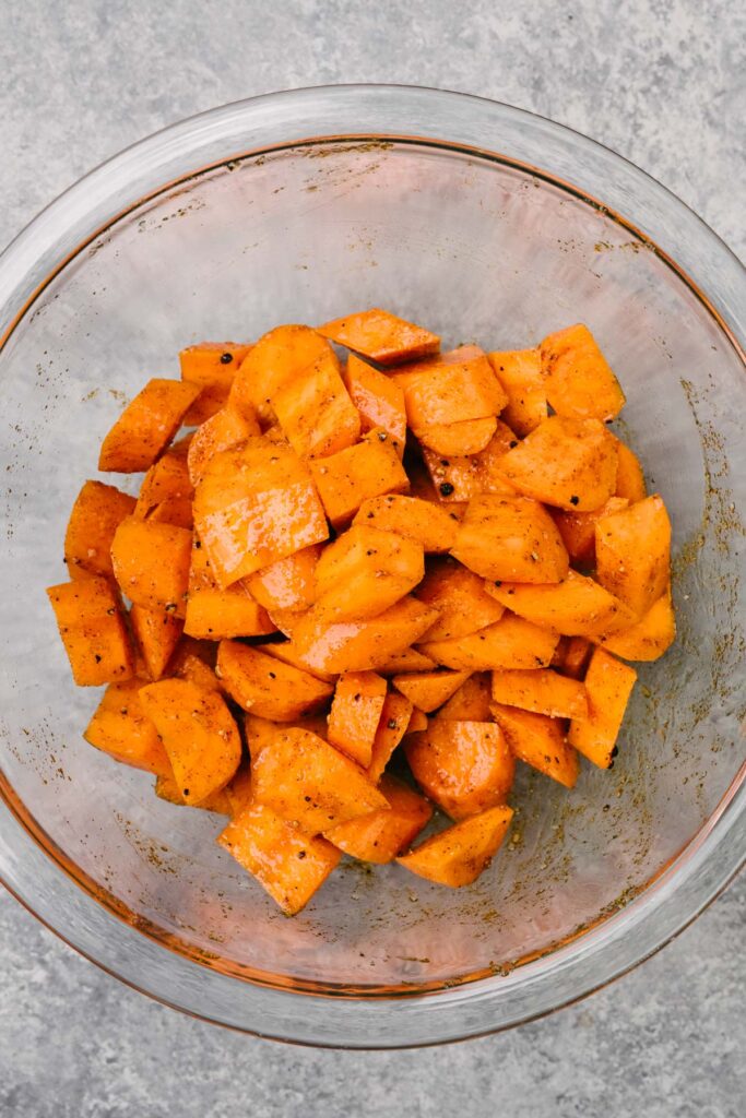 Sliced carrots tossed with olive oil, salt, pepper, cumin, paprika, and garlic powder in a glass mixing bowl.