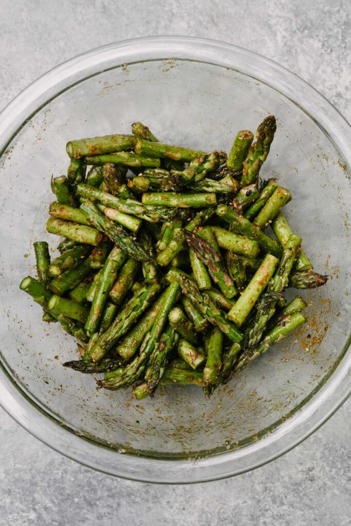 Chopped asparagus tossed with olive oil, salt, pepper, cumin, paprika, and garlic powder in a glass mixing bowl.