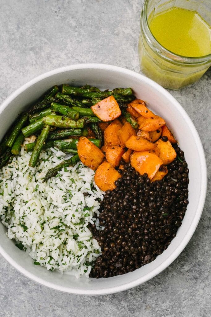 The base of a mediterranean bowl in a low white bowl with a small jar of lemon vinaigrette to the side; the base consists of herb white rice, black lentils, and roasted carrots and asparagus.