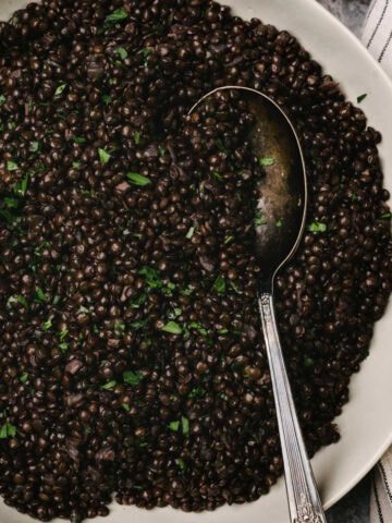 A silver serving spoon tucked cooked black lentils in a shallow serving bowl, garnished with fresh parsley; a striped linen napkin is tucked to the side.