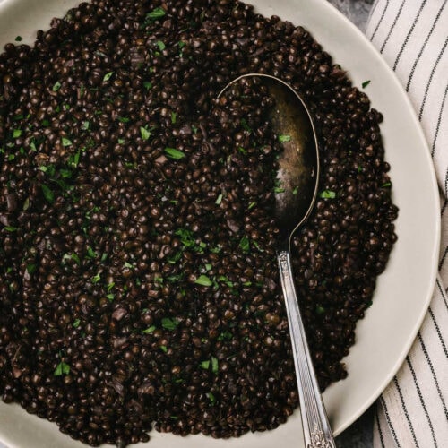 A silver serving spoon tucked cooked black lentils in a shallow serving bowl, garnished with fresh parsley; a striped linen napkin is tucked to the side.