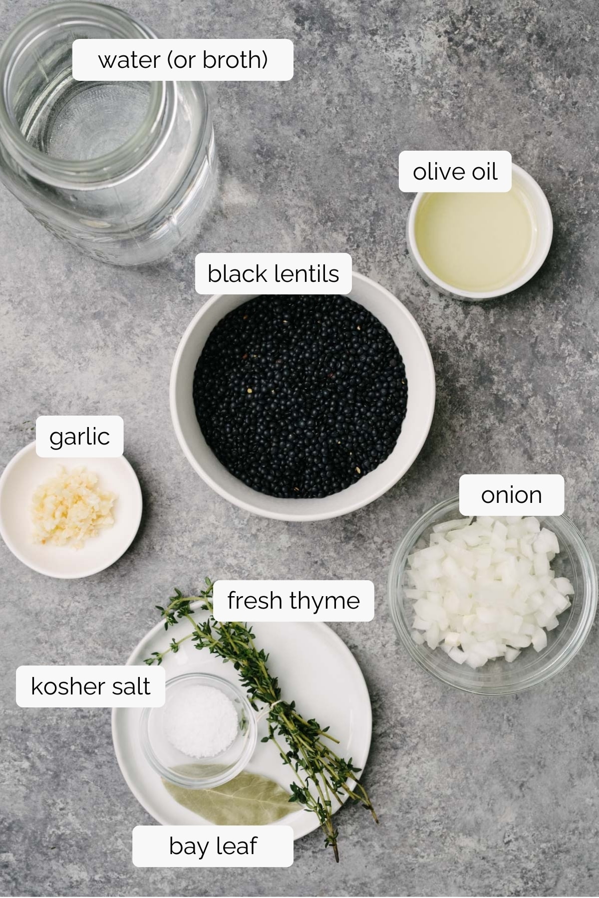 The ingredients to cook black lentils in small bowls arranged on a concrete background - dried lentils, olive oil, onion, garlic, dried lentils, water, thyme, bay leaf, and salt.