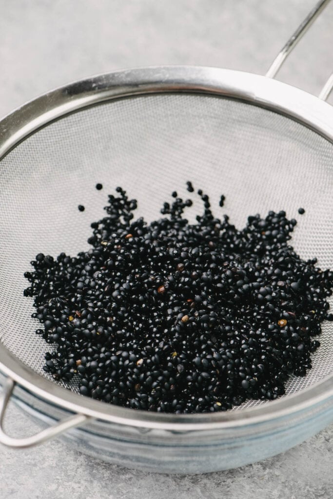 Side view, dried black beluga lentils in a mesh strainer nested in a glass bowl.