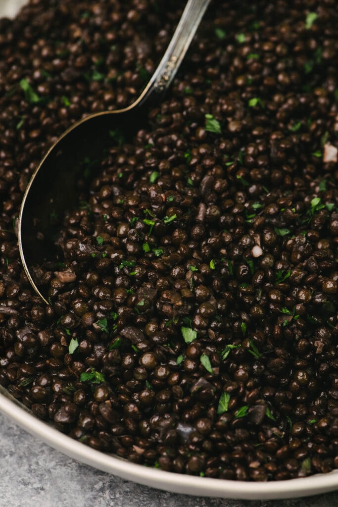 Side view, a spoon tucked into cooked black lentils seasoned with salt, lemon juice, and fresh parsley in a tan serving bowl.