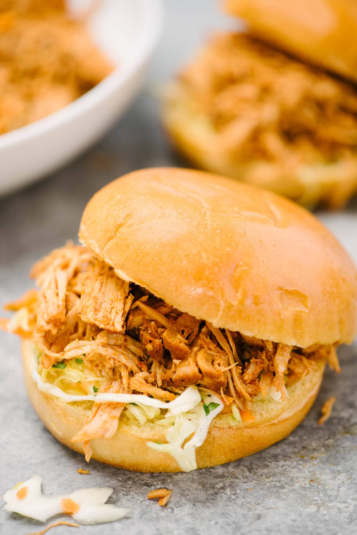 Side view, slow cooker pulled pork sandwich with coleslaw on a brioche bun; a serving bowl of pulled pork and a second sandwich are in the background.