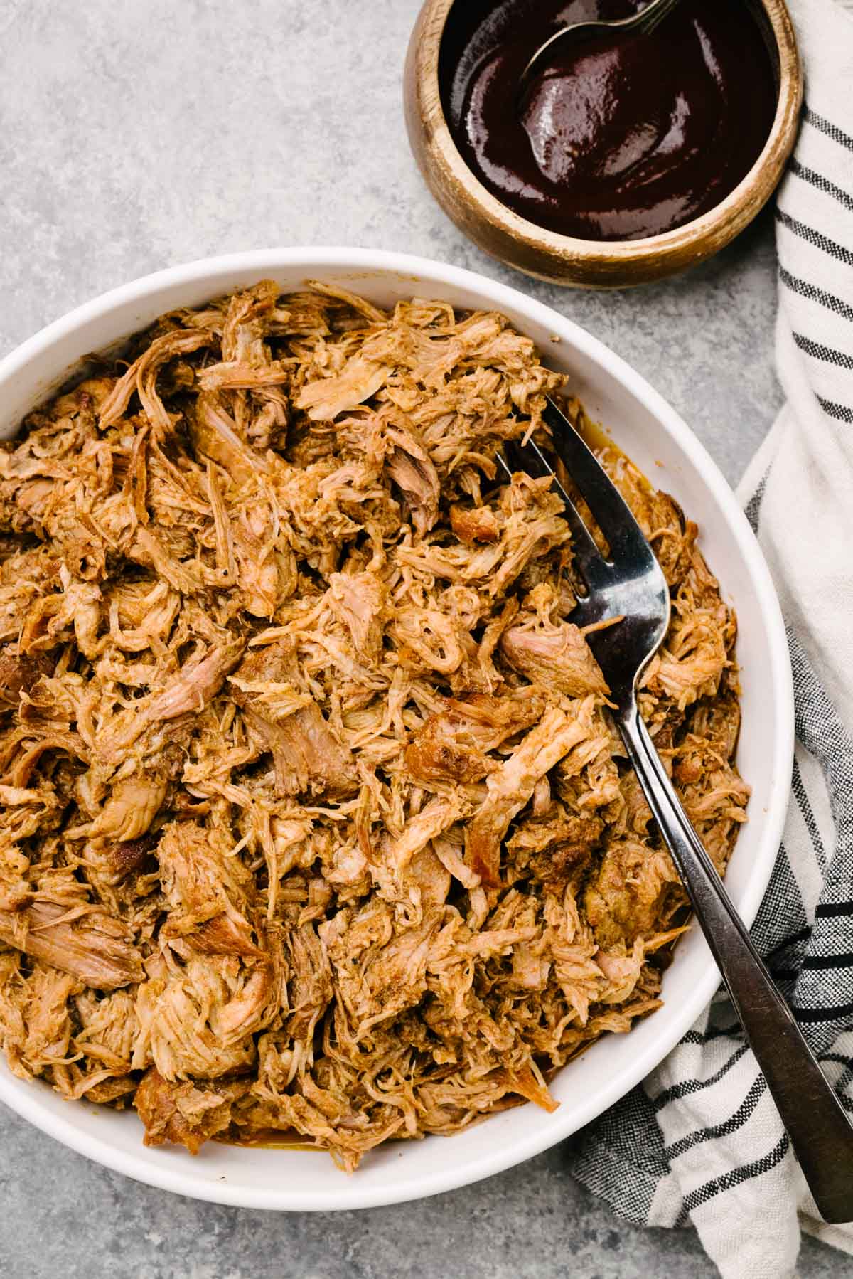 A serving fork tucked into a large white bowl of crockpot pulled pork; a bowl of barbecue sauce and striped linen napkin surround the bowl.