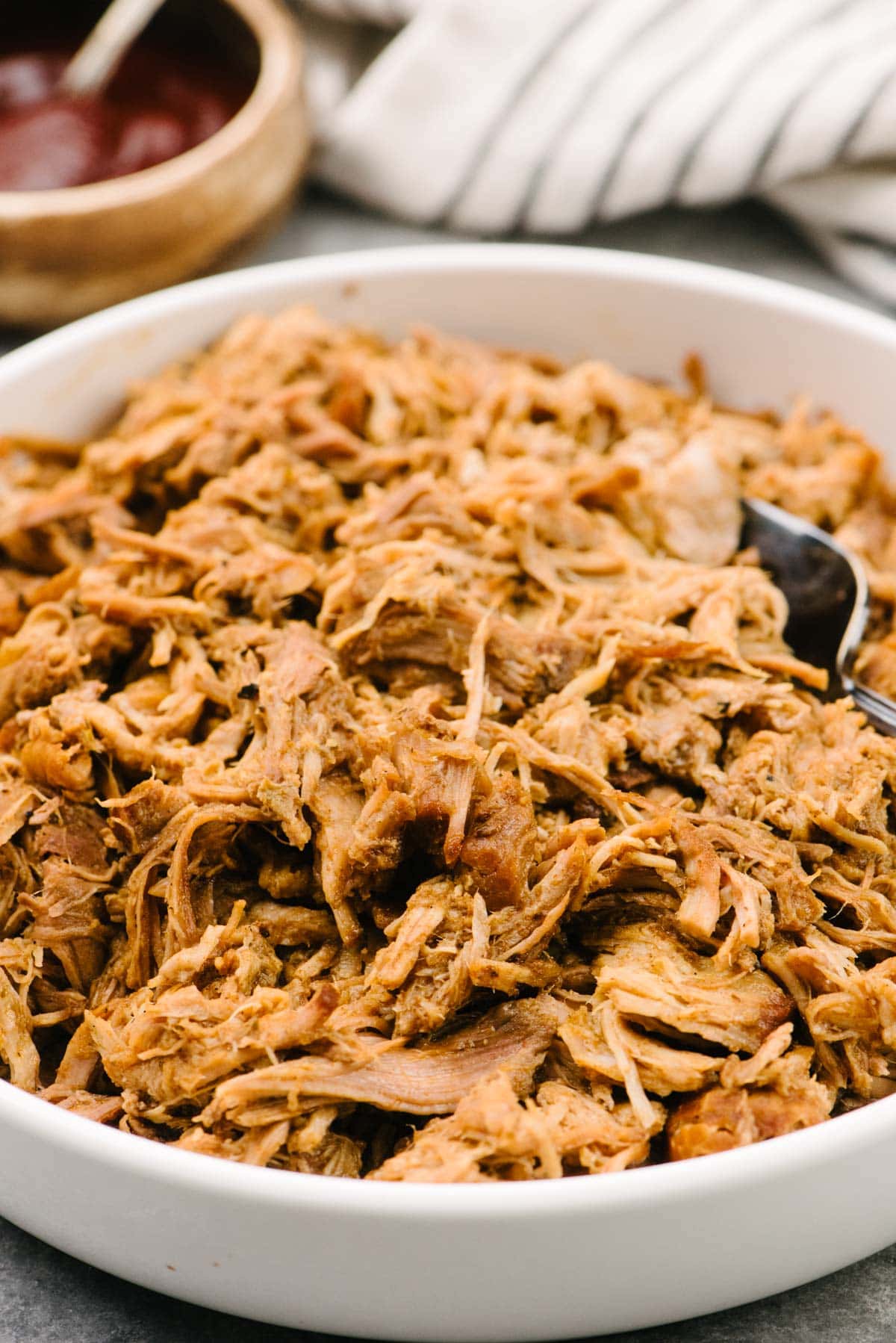 Side view, a serving fork tucked into a bowl of slow cooker pulled pork with a bowl of barbecue sauce and striped linen napkin the background.