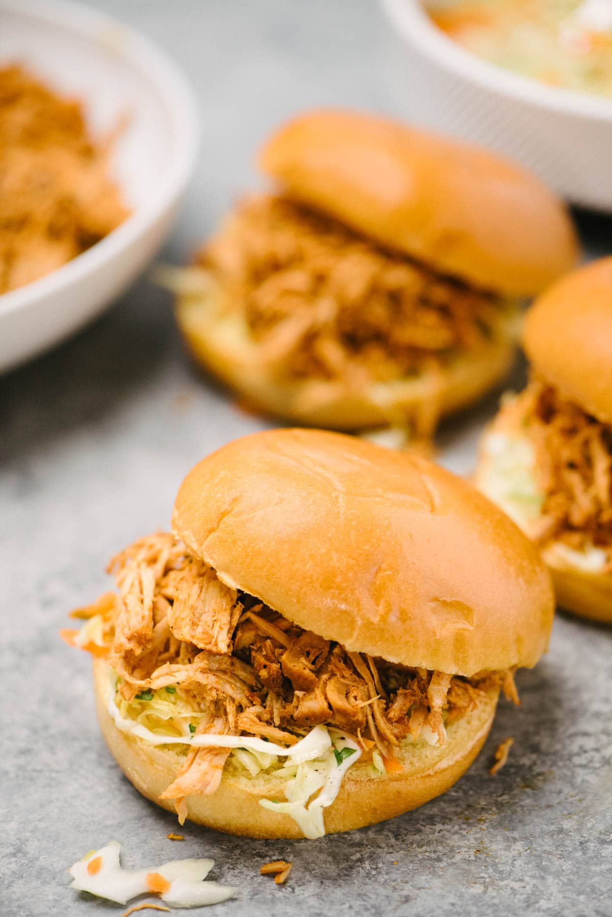 Side view, three pulled pork sandwiches with coleslaw on brioche buns; bowls of pulled pork and coleslaw are in the background.