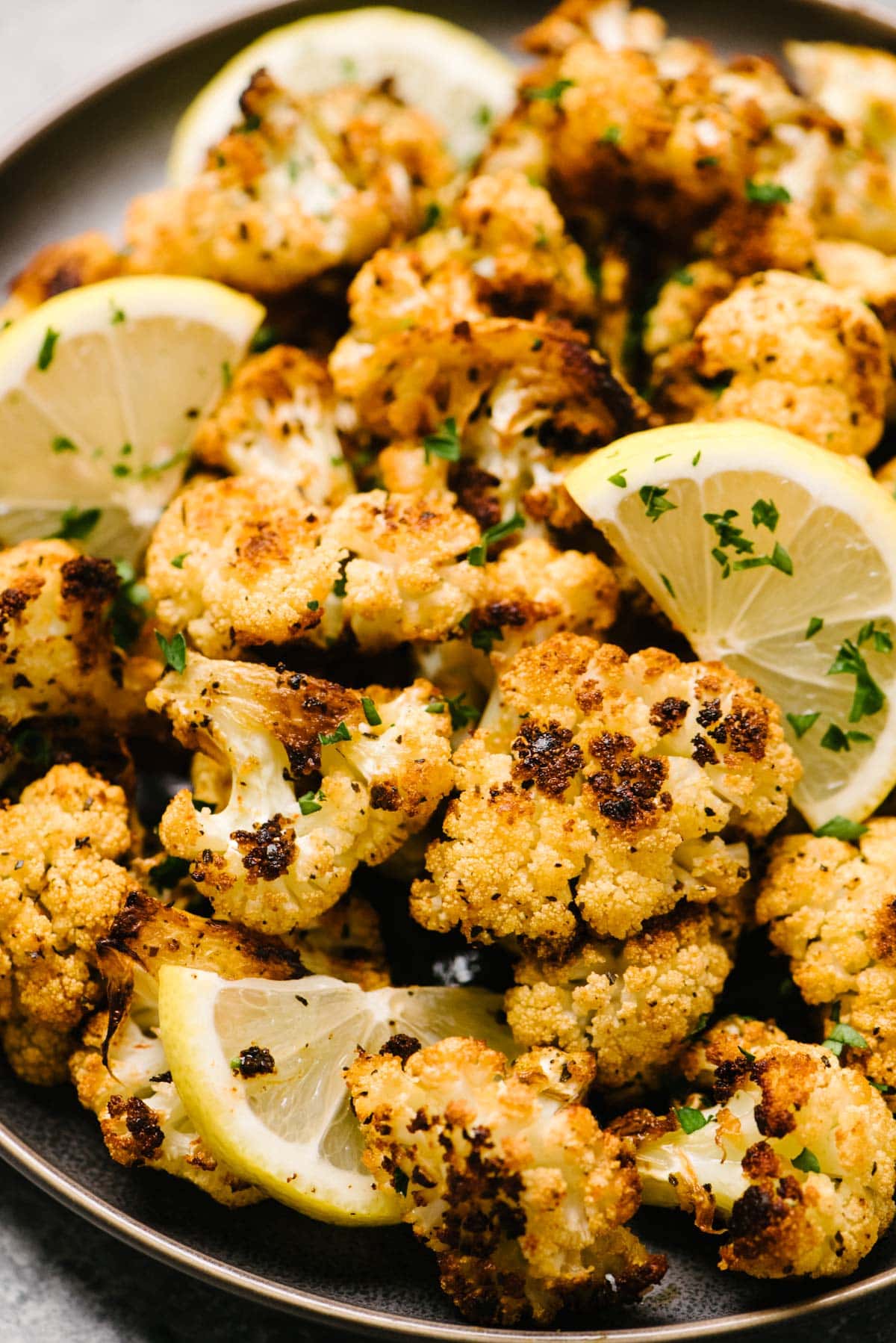 Side view, roasted cauliflower on a black plate garnished with lemon wedges and fresh minced parsley.