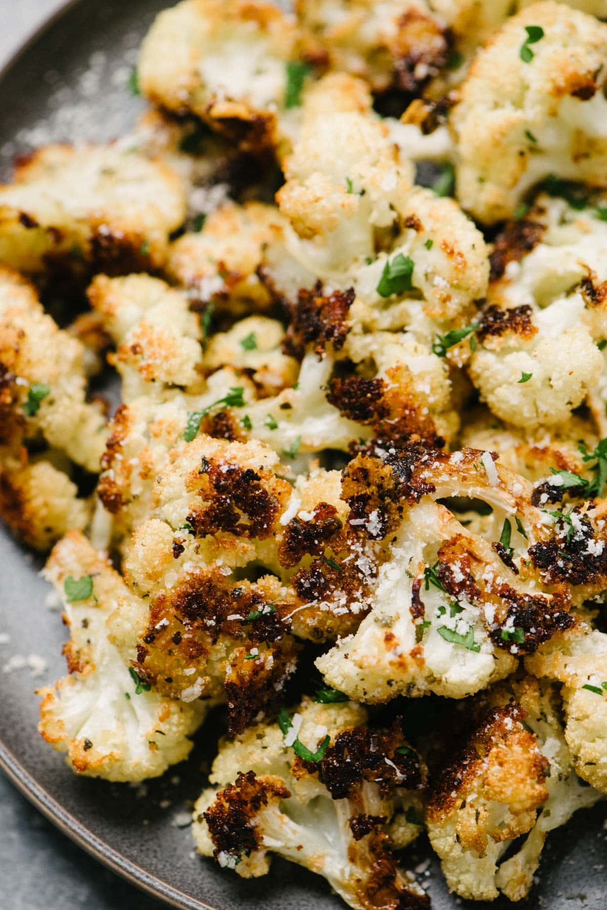 Side view, roasted cauliflower with grated parmesan cheese and finely chopped fresh basil on a dark grey plate.