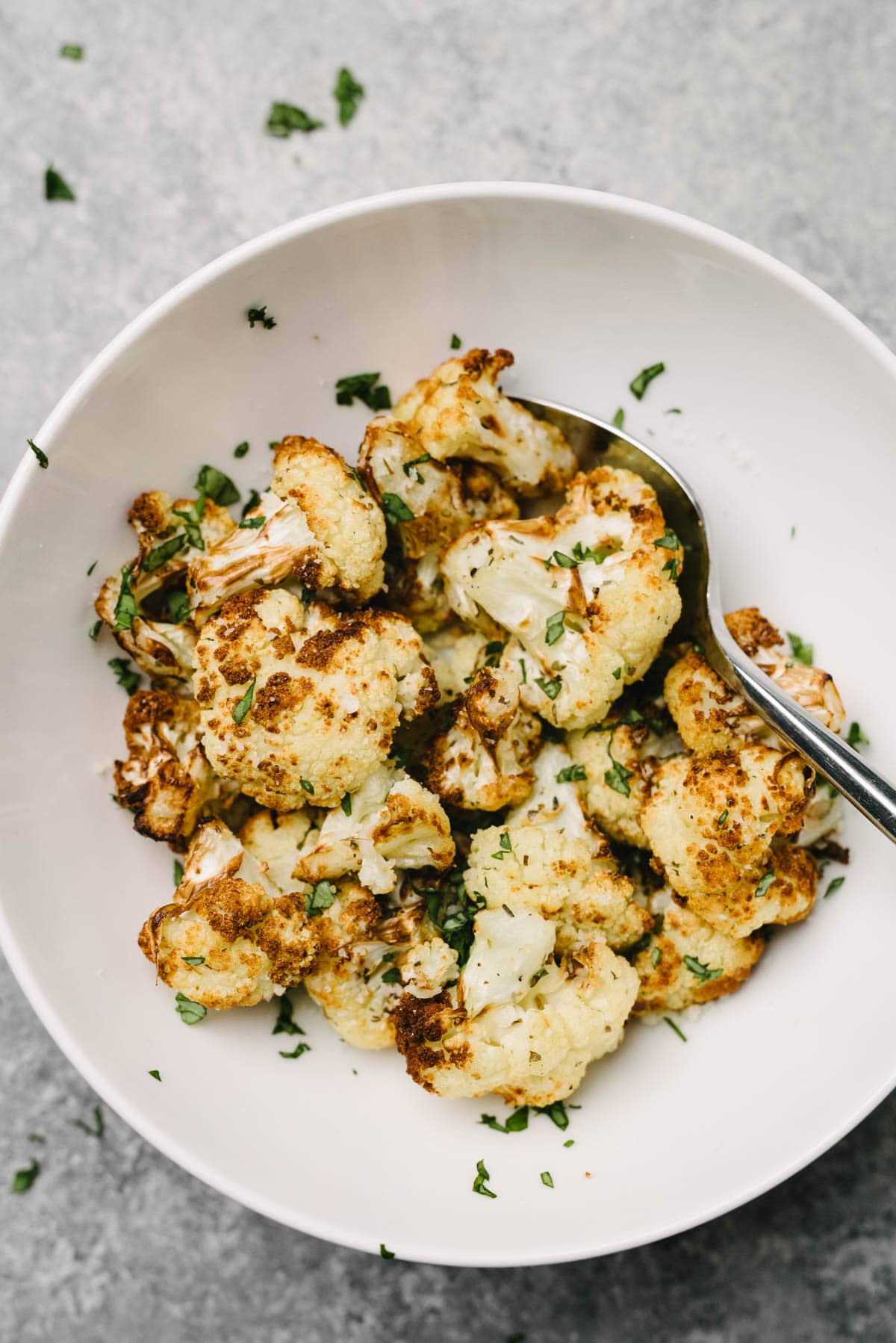 A spoon tucked into a bowl of roasted cauliflower florets, garnished with fresh chopped basil.