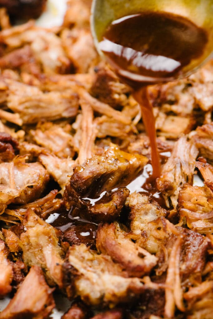 Side view, drizzling the reduced pulled pork pan sauce over crispy shredded pulled pork on a baking sheet.