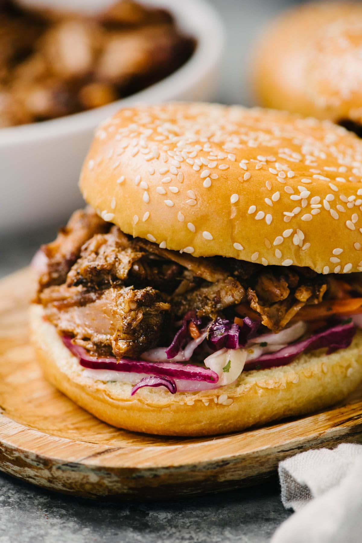 Side view, a pulled pork sandwich with coleslaw on a brioche bun on a wood serving board; a bowl of pulled pork and a second sandwich are in the background.