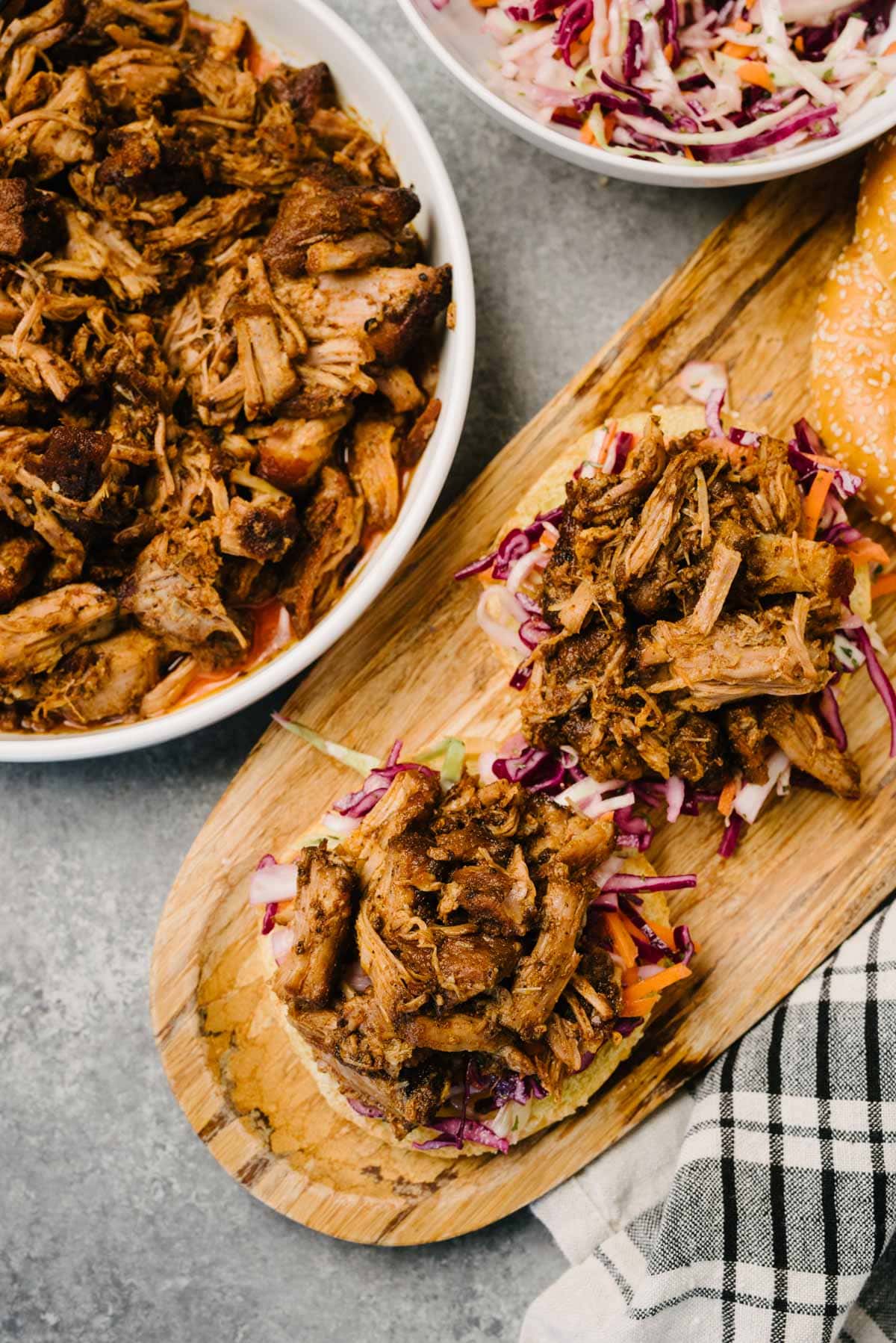 Two buns topped with coleslaw and pulled pork on a wood serving board. The board is surrounded by a bowl of pulled pork, a bowl of coleslaw, and a linen napkin.