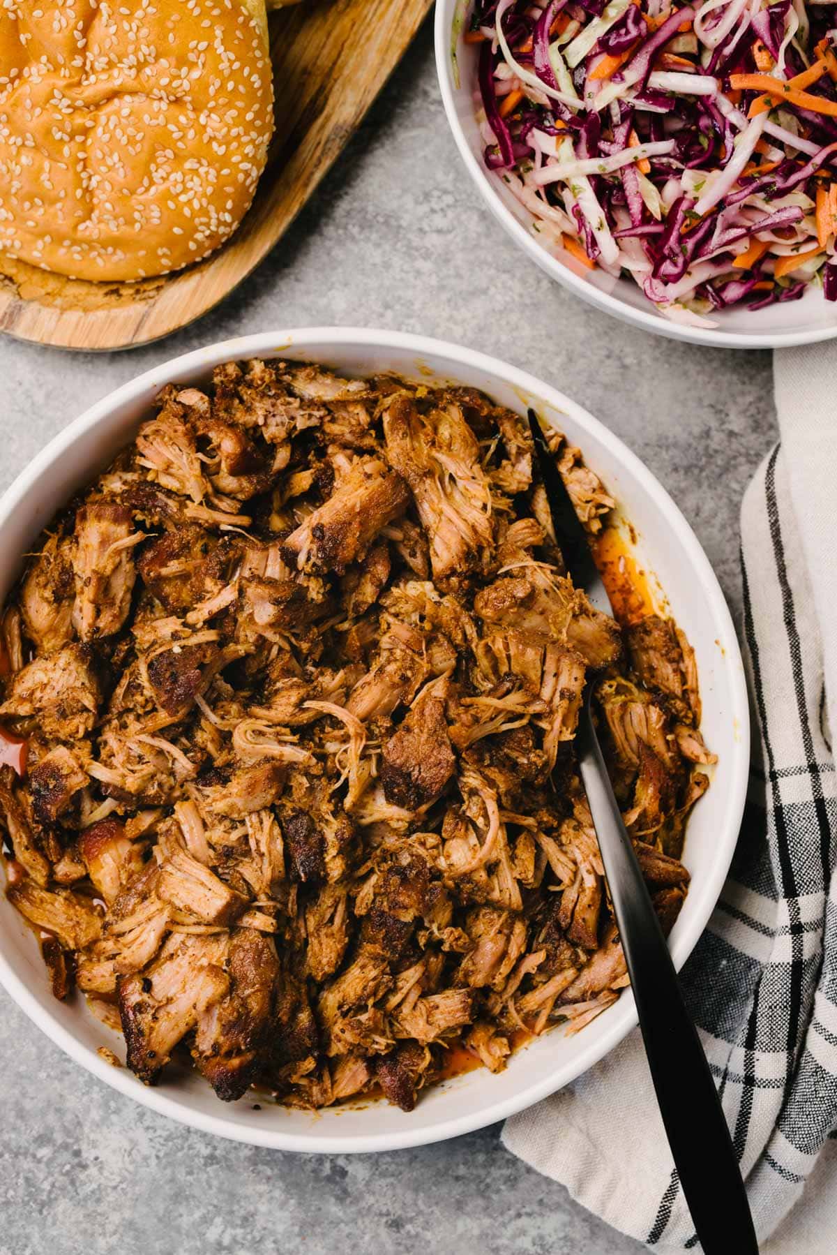 A black serving fork tucked into a white serving bowl of pulled pork; a bowl of coleslaw, tray of brioche buns, and a linen napkin surround the bowl.
