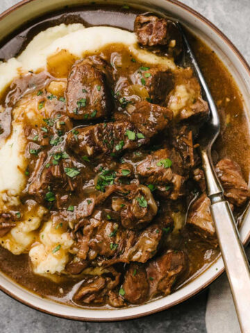 A fork tucked into tender Instant Pot beef tips over mashed potatoes in a low shallow bowl, with a striped linen napkin to the side.