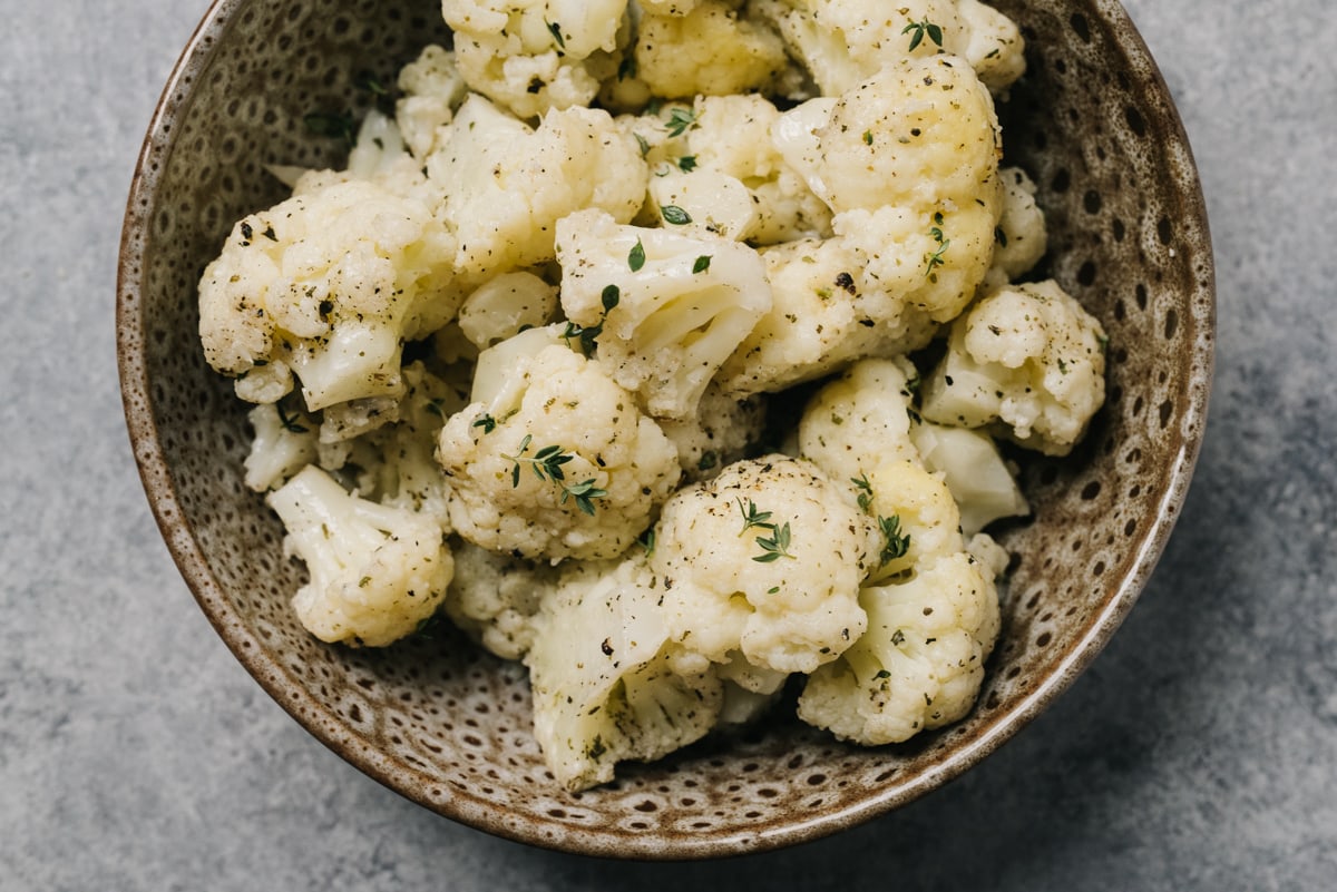Instant pot cauliflower florets tossed with olive oil, salt, pepper, and fresh thyme in a brown speckled serving bowl.