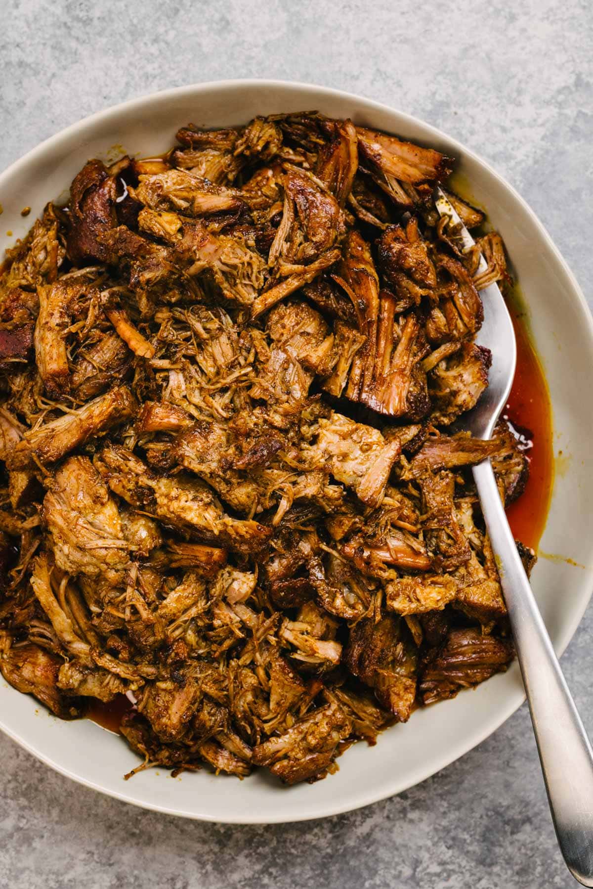 A silver serving fork tucked into a bowl of Instant Pot pulled pork, drizzled with pan sauce.