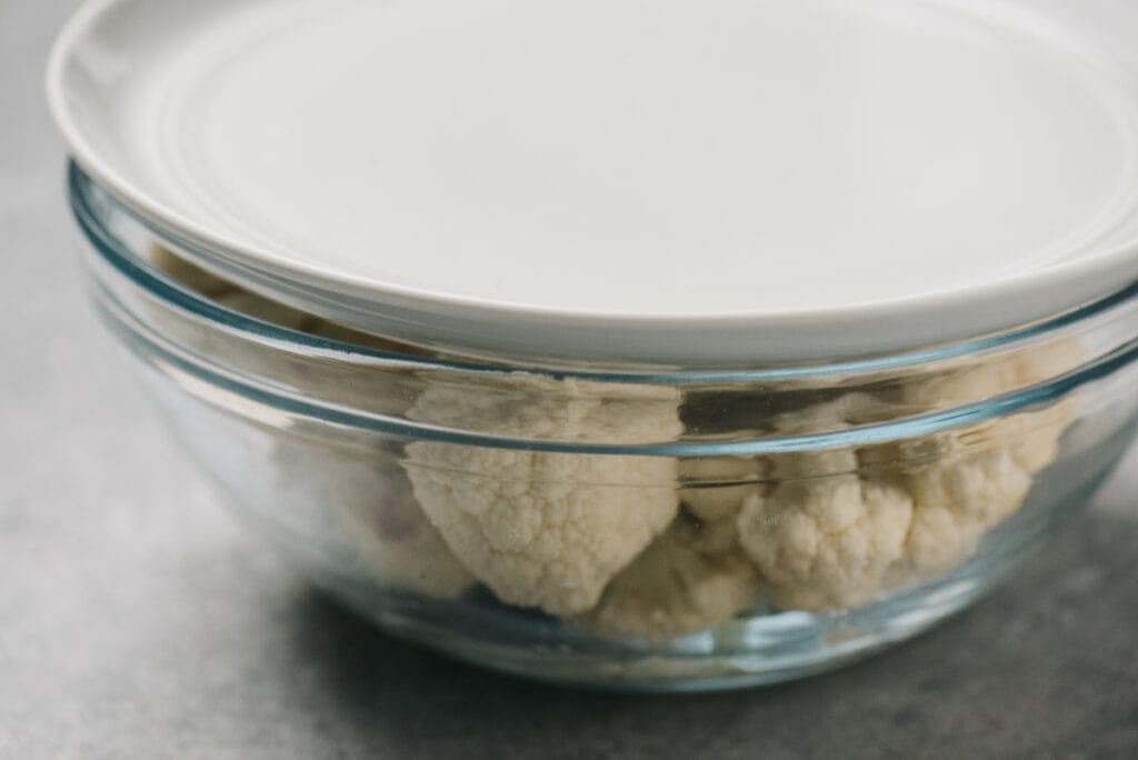 Side view, cauliflower florets with water in a glass mixing bowl topped with a plate to trap in steam.