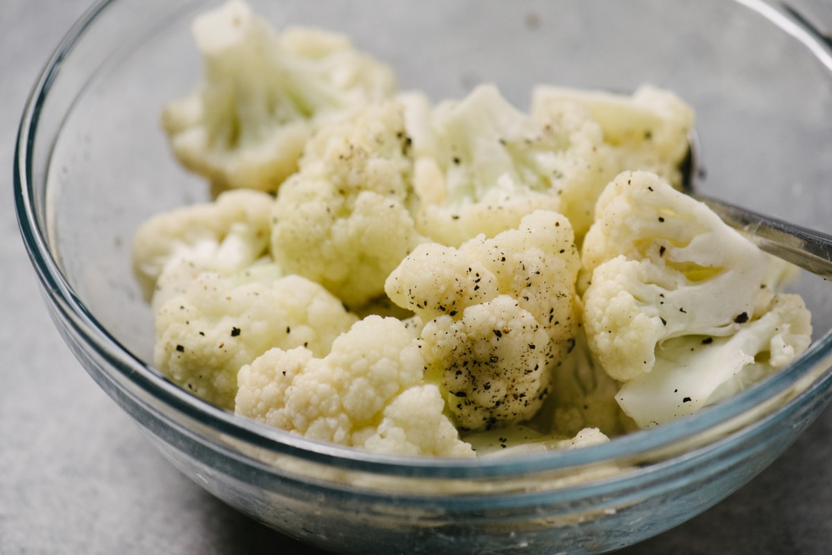 Side view, microwave steamed cauliflower in a glass mixing bowl tossed with butter, salt, and pepper.
