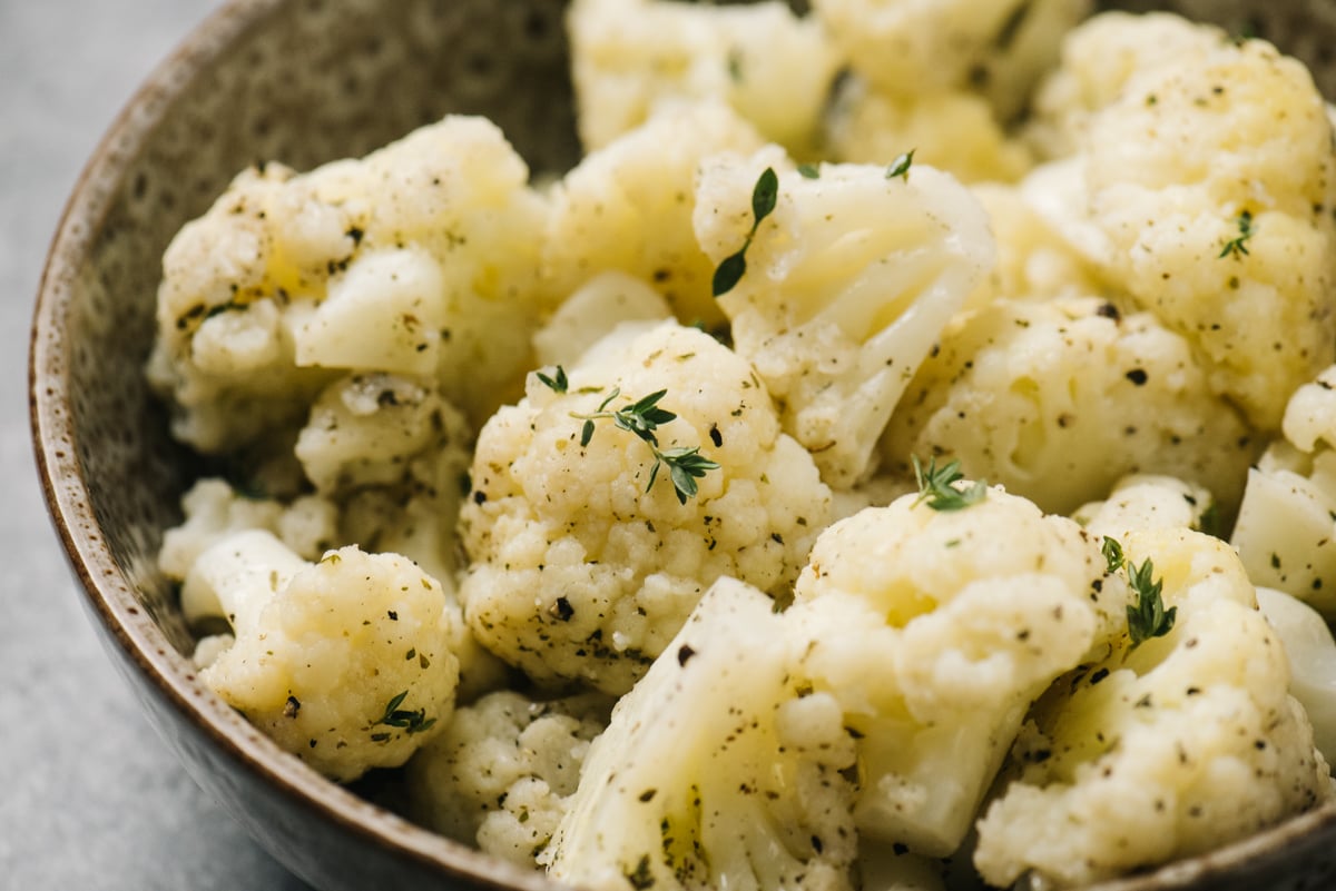 Side view, steamed cauliflower tossed with olive oil, salt, pepper, and fresh thyme in a brown speckled serving bowl.
