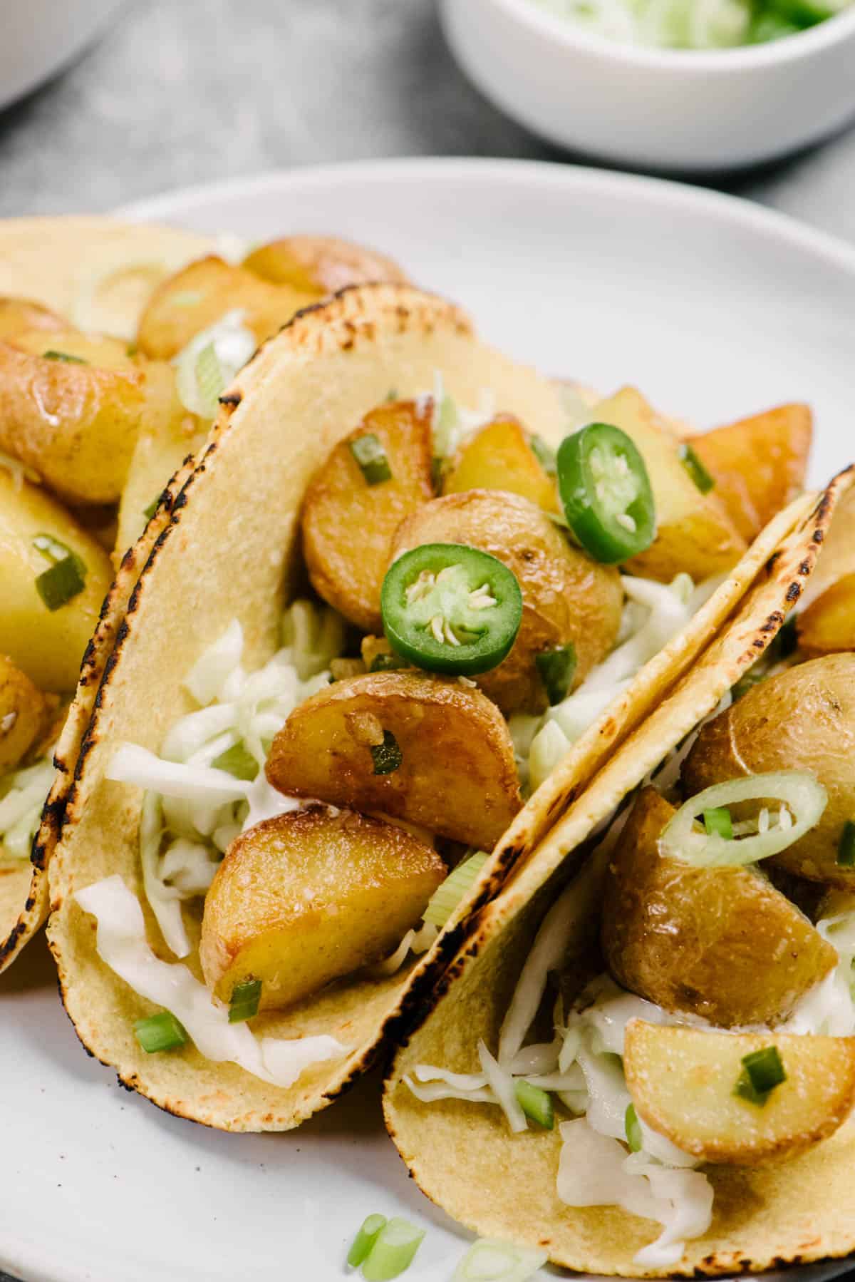 Side view, three potato tacos with serrano chilies on a white plate, garnished with coleslaw and green onions.