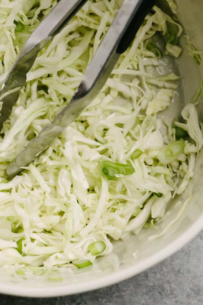 A pair of tongs tucked into a bowl of lime coleslaw, made with cabbage, green onions, and lime juice.