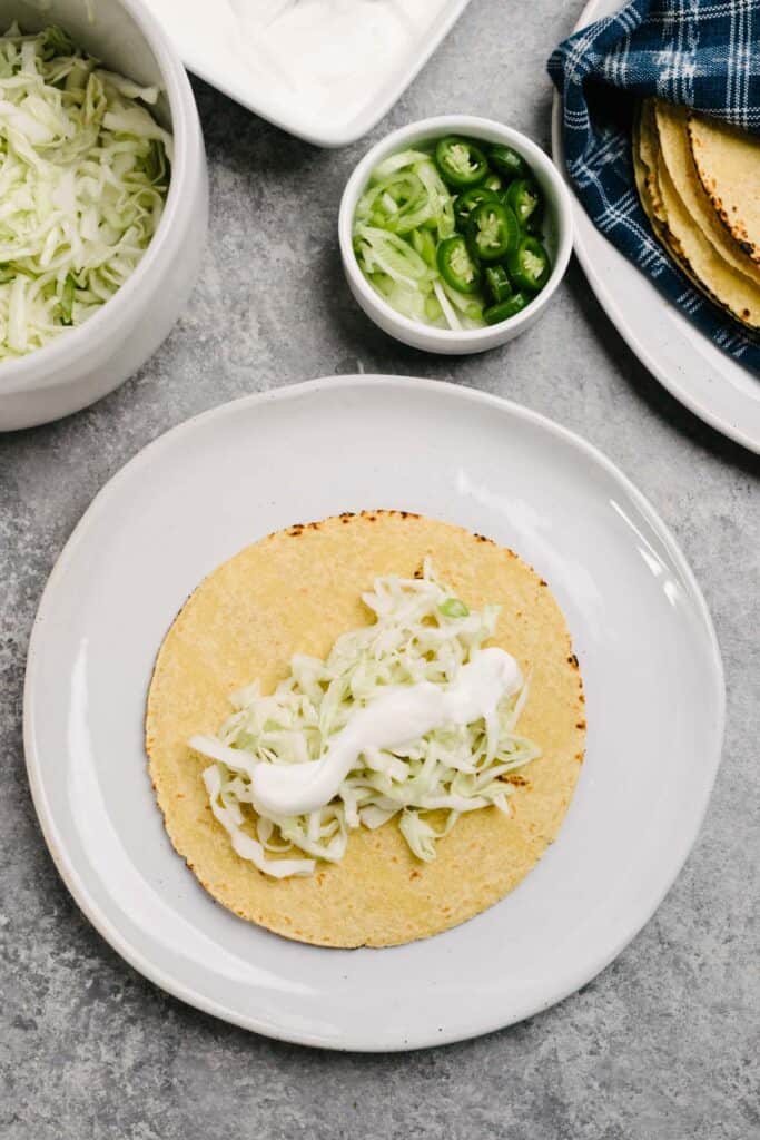 A corn tortilla layered with lime coleslaw and sour cream on a white plate.